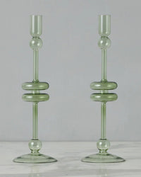 The etúHOME Sage Glass Candlesticks Set of 2 - Large, crafted from recycled glass in translucent green with round and disc accents, stand side by side on a white marble surface.