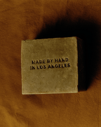 A rectangular Manuka Honey Bar Soap by Flamingo Estate, stamped with MADE BY HAND IN LOS ANGELES, rests on a brown fabric background.