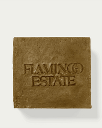 A square, brown Manuka Honey Bar Soap by Flamingo Estate is embossed with the brand’s name in capitals, set against a light background that highlights its luxurious and hydrating skincare benefits.