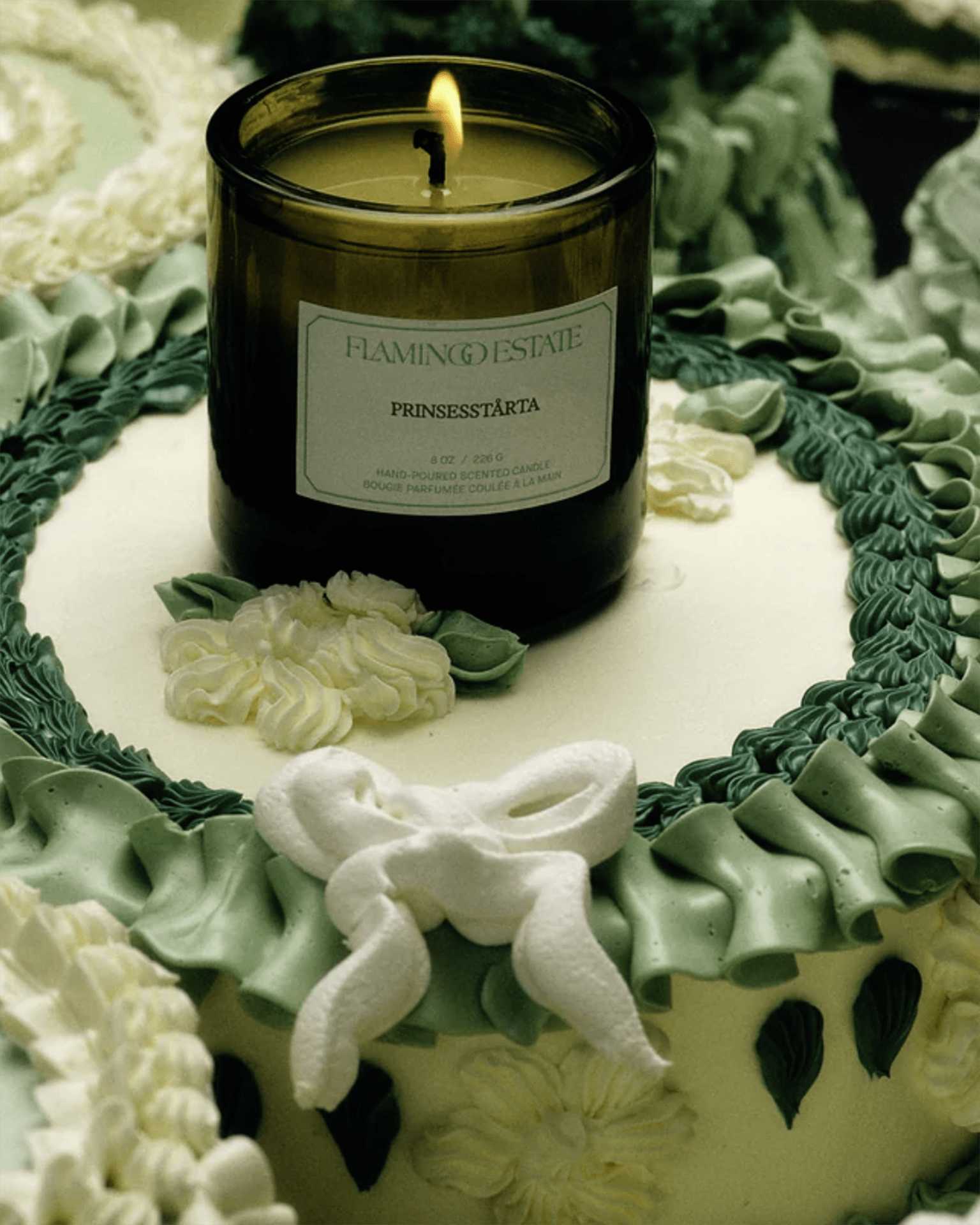 A green and white cake with icing flowers and a bow, topped with a lit Flamingo Estate Prinsesstarta Candle in a green glass jar.