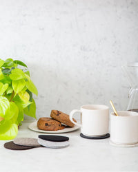 Two white mugs, a gold spoon, cookies on a plate, a green plant, and a glass coffee maker sit on a white marble countertop alongside graf lantz Bierfilzl Merino Wool Felt Round Coaster Multi in Earth—sustainable home décor.