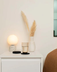 A white nightstand features a round lamp, a glass, graf lantz Bierfilzl Merino Wool Felt Round Coasters in black, and a white vase with dried pampas grass, beside a bed topped with a brown blanket.