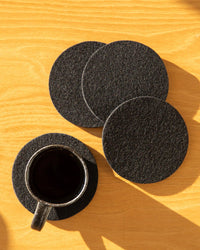 A glass cup of black coffee rests on a graf lantz Bierfilzl Merino Wool Felt Round Coaster in black, with three more coasters from the 4-pack set nearby, all arranged on a light wooden surface.