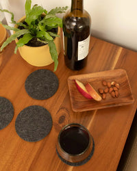 A wooden table with a potted plant, wine bottle, glass of red wine on graf lantz Bierfilzl Merino Wool Felt Round Coaster Solid in Charcoal (4 Pack), plus a tray with apple slices and almonds—perfect sustainable home decor.