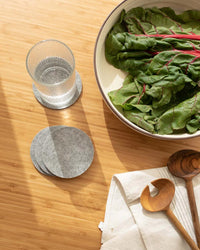 A bowl of leafy greens and a glass of water rest on a wooden surface with graf lantz Bierfilzl Merino Wool Felt Round Coaster in Granite (4-Pack), plus extra sustainable coasters and wooden utensils on a napkin.