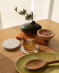 A glass of amber liquid rests on a graf lantz Bierfilzl Merino Wool Felt Round Coaster in Heather White, paired with a green plate, wooden spoon, pink salt bowl, vase with branch, and stacked books for an inviting sustainable table decor.