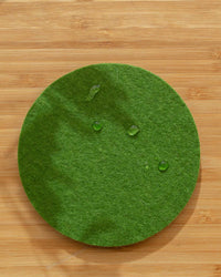 A graf lantz Bierfilzl Merino Wool Felt Round Coaster in Loden Green from the 4 Pack sits on a light wooden surface, with three small water droplets visible on it.