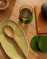 A wooden spoon rests on a yellow plate beside a glass of sparkling drink on a graf lantz Bierfilzl Merino Wool Felt Round Coaster in Loden Green, an orange notebook, and a bowl of pink salt atop a wooden surface.