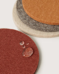 The Bierfilzl Merino Wool Round Coaster Multi in Mesa - 4 Pack by graf lantz features four sustainable German Merino wool coasters in red, beige, gray, and orange. Water beads atop the red coaster, highlighting their protective design.