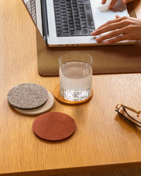 A hand types on a laptop at a wooden desk with a glass of water resting on graf lantz Bierfilzl Merino Wool Round Coasters Multi in Mesa (4 Pack), felt coasters, and eyeglasses nearby.