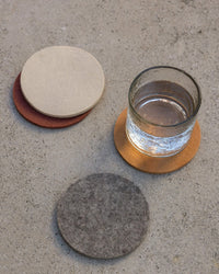 A glass of water rests on a graf lantz Bierfilzl Merino Wool Round Coaster Multi in Mesa (4 Pack), with grey and beige coasters beside it, all on concrete—a stylish, sustainable décor accent.
