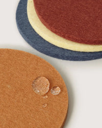 The graf lantz Bierfilzl Merino Wool Round Coaster Multi in Yucca - 4 Pack features brown, blue, yellow, and red sustainable coasters stacked, with water droplets on the brown coaster in front.