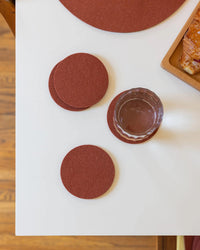 A glass of water rests on a graf lantz Bierfilzl Merino Wool Round Coaster in Terracotta, with three more from the 4-pack nearby on a white table—a stylish, sustainable addition to your home décor.