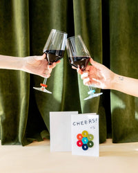 Two hands clink red wine glasses before green curtains. On the table, a graf lantz Cheers Wine-Otes Merino Wool Felt Wine Marker Note Card and playful wine charms add a festive touch to the celebration.