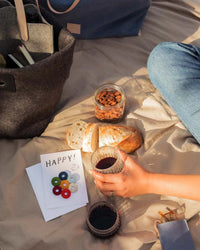 At a picnic, a person holds a glass of wine on a blanket with bread, almonds, and graf lantzs Happy Wine-Otes Merino Wool Felt Wine Marker Note Card that reads HAPPY!, along with colorful wine charms and other items.