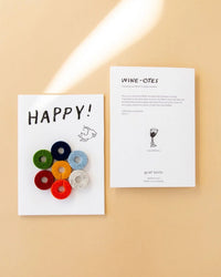The graf lantz Happy Wine-Otes Merino Wool Felt Wine Marker Note Card features “HAPPY!” text and seven colorful felt circles, displayed with an instruction card on a beige background.