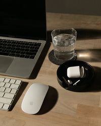 A laptop, wireless mouse, keyboard, glass of water, and wireless earbuds in a charging case are neatly arranged on a wooden desk with the graf lantz Kobon Leather Round Tray in Black for added organization.