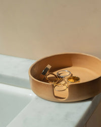 The graf lantz Kobon Leather Round Tray in Natural organizes metal rings and a bracelet, resting elegantly on the edge of a white marble countertop.