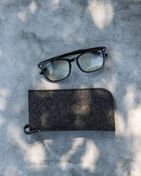 Black eyeglasses and the graf lantz Merino Wool Felt Anzen Eyeglass Sleeve in Charcoal rest on light grey concrete, with sunlight and shadows creating an elegant look while offering superior eyewear protection.