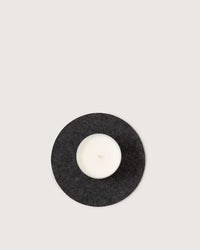 A white candle in a glass holder rests on the graf lantz Round Merino Wool Felt Trivet 8 in Charcoal—an elegant, sustainable kitchen accessory.