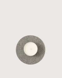 A white candle in a glass holder rests on the graf lantz Round Merino Wool Felt Trivet 8 in Granite, a sustainable kitchen accessory, against a plain light background.