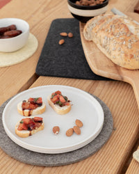 A plate with three pieces of bruschetta and two almonds sits beside a cutting board with seeded bread, a bowl of dates, and the graf lantz Round Merino Wool Felt Trivet 8 in Granite—an eco-friendly kitchen accessory.