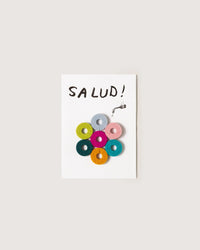 The graf lantz Salud Wine-Otes Merino Wool Felt Wine Marker Note Card displays SALUD! above a faucet with seven colorful wine charms in a hexagon—perfect as a playful host gift.