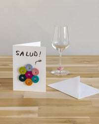 The graf lantz Salud Wine-Otes Merino Wool Felt Wine Marker Note Card features colorful felt wine charms and a festive SALUD! message—an ideal host gift placed beside a glass, envelope, and table.