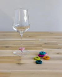 A graf lantz Salud Wine-Otes Merino Wool Felt Wine Marker Note Card rests on a wooden table beside colorful felt rings, perfect as unique wine charms or a thoughtful host gift.