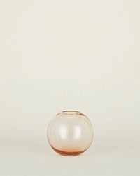 The Aurora Sphere Vase in Blush by Hawkins New York, a small round vessel of translucent pink handblown glass, rests on a plain light-colored surface against a neutral background.