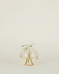 The Aurora Sphere Vase in Blush by Hawkins New York features handblown transparent glass with pale pink tones, holding delicate white flowers and green stems against a simple light background.