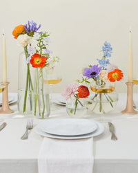 A table set with white plates, cutlery, and napkins features floral arrangements in Hawkins New York Aurora Sphere Vases in Clear and two tall candles in holders.