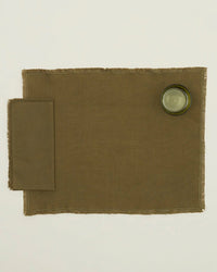 An Essential Dinner Napkin in Olive by Hawkins New York is placed on the left of a brown placemat, with a clear drinking glass at the top right, all set against a plain white background.