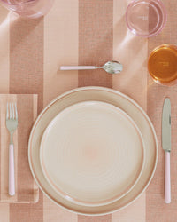 A place setting with a fork, knife, and spoon with pink handles, Hawkins New York Essential Dinner Plates in Blush, and pink and amber glasses on a peach striped tablecloth.
