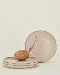 A sweet potato with a purple sprout rests on a stack of three blush Essential Dinner Plates by Hawkins New York, with a fourth plate upright in the background. This elegant stoneware set of 4 is dishwasher safe.