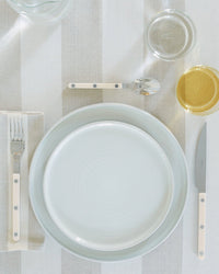 A neatly arranged table setting features Hawkins New York Essential Dinner Plates in Bone (set of 4), a fork, knife, spoon, two plates, a water glass, and a yellow drink on a striped tablecloth.