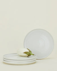 A white eggplant sits on a stack of three Hawkins New York Essential Dinner Plates in Bone (set of 4), with the fourth stoneware plate standing upright against a plain light background.