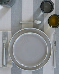 A table setting with a fork, knife, spoon, glass, cup, napkin, gray-striped tablecloth, and two stacked Essential Dinner Plates in Light Grey from Hawkins New York.
