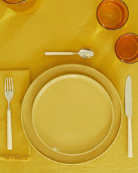 A table setting with an Essential Dinner Plate in Mustard from Hawkins New York (set of 4), on a yellow tablecloth, paired with white-handled cutlery, a yellow napkin, and two amber drinking glasses.