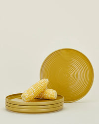 Three Essential Dinner Plates in Mustard from Hawkins New York are stacked with two ears of corn on top, while a fourth plate from the set of 4 stands upright in the background against a plain backdrop.