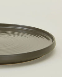 A close-up of a round Hawkins New York Essential Dinner Plate in Olive, showing its dark stoneware surface with a slightly raised rim and circular patterns; available as a set of 4.