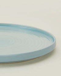 A close-up of a round, sky blue Essential Dinner Plate by Hawkins New York, featuring a slightly raised rim, set against a plain off-white background. Part of the Essential Dinner Plates in Sky - Set of 4.