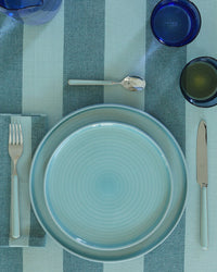 A place setting with a blue-striped tablecloth, Hawkins New York Essential Dinner Plates in Sky, silverware, and blue glasses arranged neatly.
