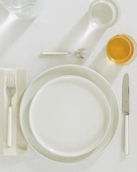 A neatly arranged place setting displays Hawkins New Yorks Essential Dinnerware in Bone—16 Piece Set: two stacked white plates, fork, knife, spoon, napkin, a water glass, and an amber drink on a crisp white tablecloth.