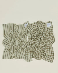 Set of two olive and sage Essential Dish Towels by Hawkins New York, each with a unique pattern—one striped, one grid—and white labels in the top right corner. Perfect absorbent cotton towels for any kitchen.