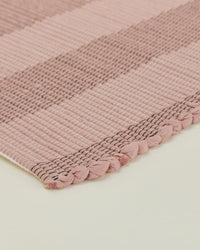 Close-up of the Hawkins New York Essential Floor Mat in Blush, featuring pink and beige stripes with braided edge detailing, displayed on a light-colored surface.