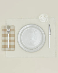 A table setting with a white plate, glass, and fork on Hawkins New York Essential Gingham Dinner Napkins in Ivory/Flax—set of 4—to the left, knife to the right, all neatly arranged on a beige placemat.