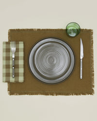 A gray ceramic plate sits on a brown placemat, flanked by a fork on Hawkins New Yorks Essential Gingham Dinner Napkins in Sage/Olive to the left, a knife to the right, and a green glass above.