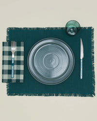 A table setting with a gray plate, fork on a Hawkins New York Essential Gingham Dinner Napkin in Sky/Peacock, knife, green glass, all arranged on a dark green fringed placemat.