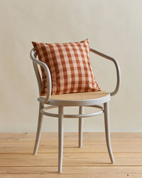 A white wooden chair with a woven seat holds the Hawkins New York Essential Gingham Pillow (18x18, Blush/Terracotta), set on a light wooden floor against a beige background.